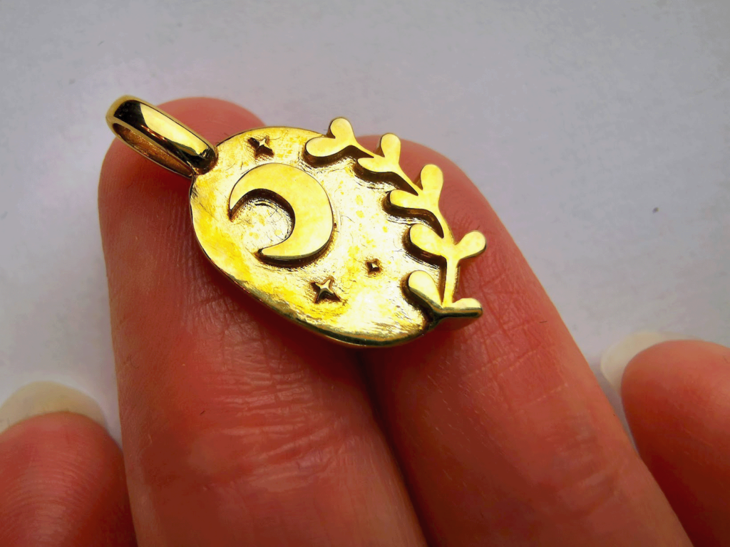 Small brass jewelry piece with a moon, three stars and a branch, presented laying on two fingers. Recessed areas are not well polished, while extruding details are shiny and smooth.